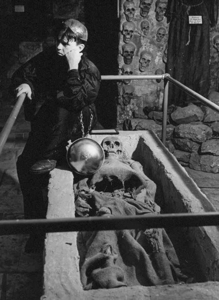 The keeper of the tombs, in the Chapel of a Thousand Skulls, sitting near a skeleton wrapped in burlap. The skeleton is known as the Guest of Honor.