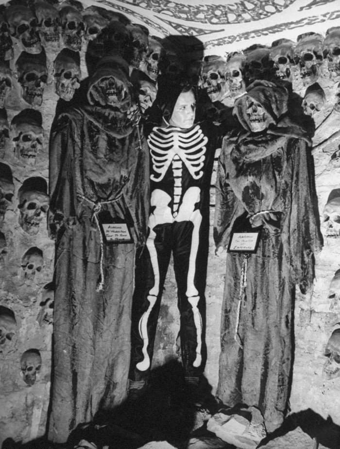 A young man, dressed as a skeleton, posing with plaster skulls and skeletons.