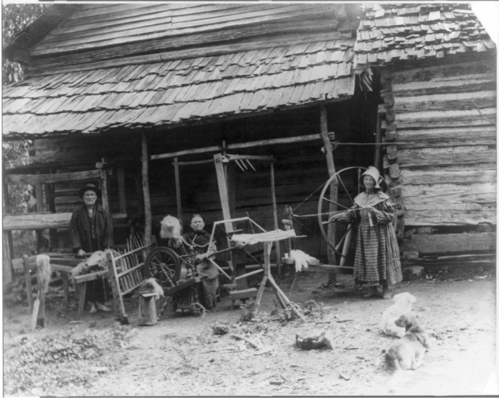Mary Faust standing next to large walking wheel, an umbrella swift, another woman seated at a spinning wheel with a distaff and a skein winder in front of her, and a man processing flax on a flax break with a counterbalance loom behind him.